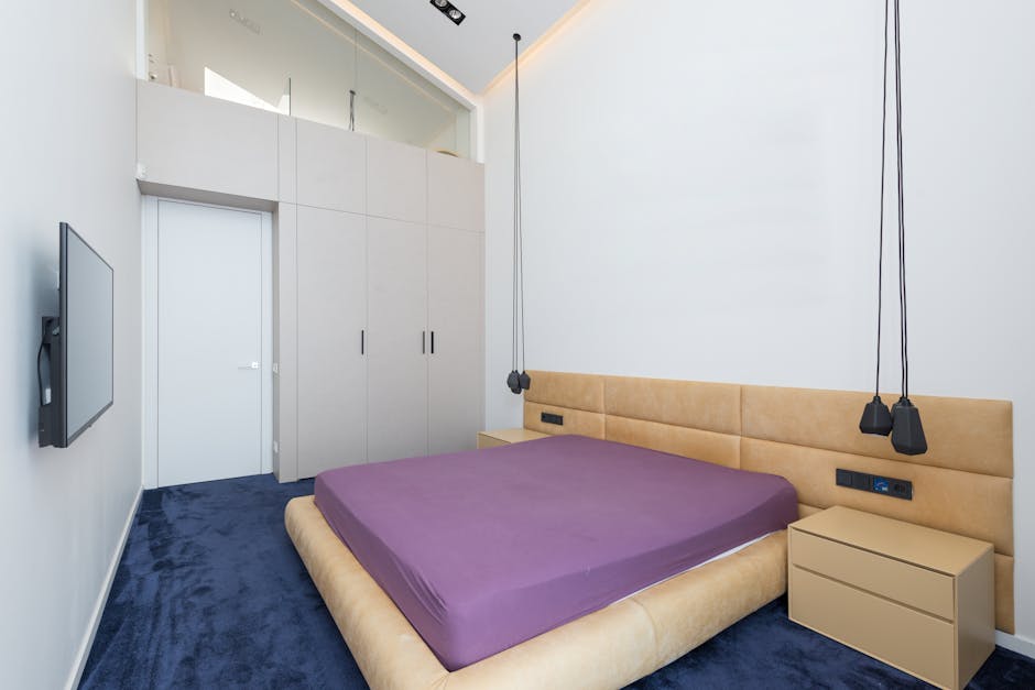 A modern bedroom with a beige upholstered bed featuring a purple mattress, positioned against a plain white wall. To the right of the bed, there is a matching beige nightstand with a smooth surface. A