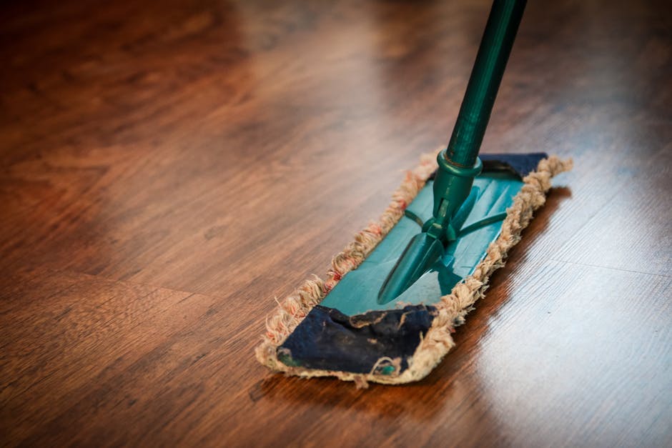 A close-up image showing a floor cleaning tool with a green handle and a rectangular cleaning head, fitted with a pad that appears used, on a wooden surface. The floor is made of polished, medium-brow
