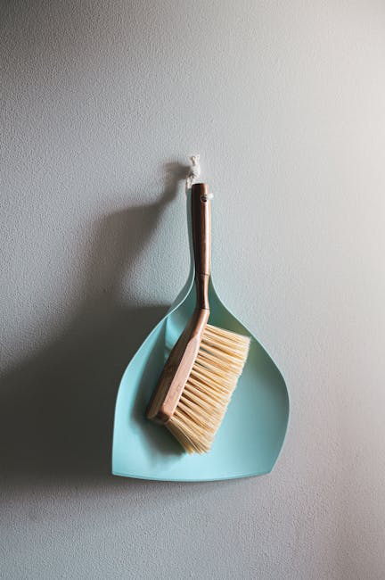 A close-up image showing a floor cleaning tool with a green handle and a rectangular cleaning head, fitted with a pad that appears used, on a wooden surface. The floor is made of polished, medium-brow